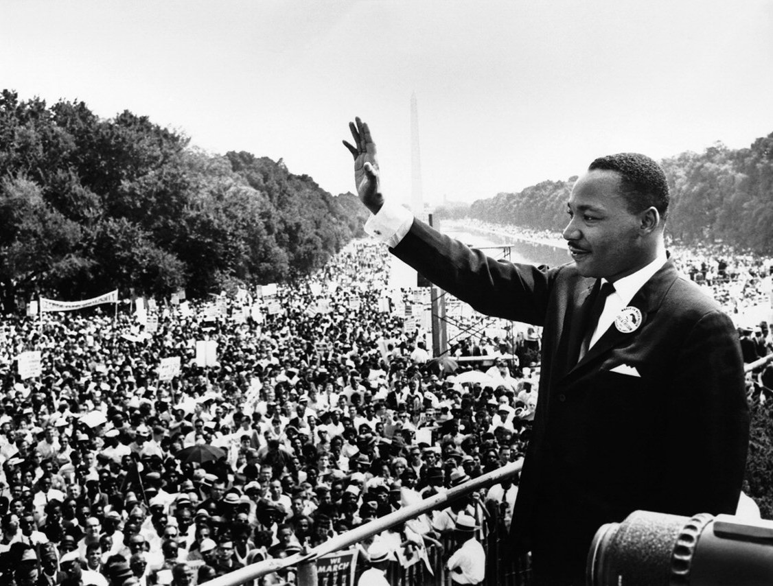 Martin Luther King raises his right hand in a wave, with thousands of people on the Washington Mall behind him